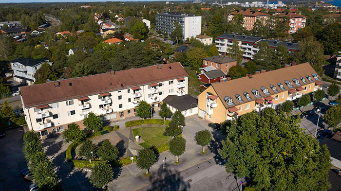 Flygfoto över Heimdalsplan i Nynäshamn, med flerfamiljshus, en innergård och omgivande grönska. Byggnaderna har röda tak och balkonger, och området är omgivet av träd och bostadshus.