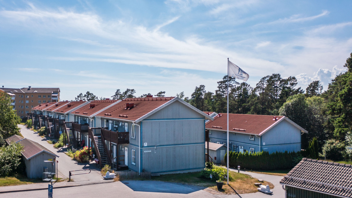 Rad av gråa radhus med röda tak längs Backluravägen 30 till 112 i Nynäshamn. Husen har utvändiga trappor och små trädgårdar. En flagga med Nynäshamnsbostäders logo vajar i förgrunden och flerfamiljshus syns i bakgrunden.