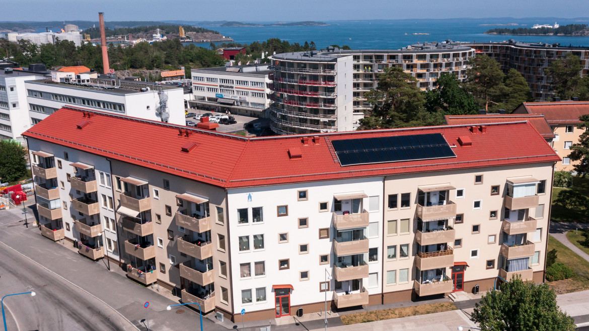 Flerfamiljshus på Nynäsvägen 12 och Industrivägen 2 i Nynäshamn, med vita fasader och röda tak. Byggnaderna har solpaneler på taket och balkonger längs fasaden. I bakgrunden syns industriområdet och skärgården.