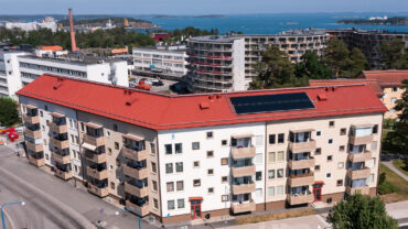 Flerfamiljshus på Nynäsvägen 12 och Industrivägen 2 i Nynäshamn, med vita fasader och röda tak. Byggnaderna har solpaneler på taket och balkonger längs fasaden. I bakgrunden syns industriområdet och skärgården.