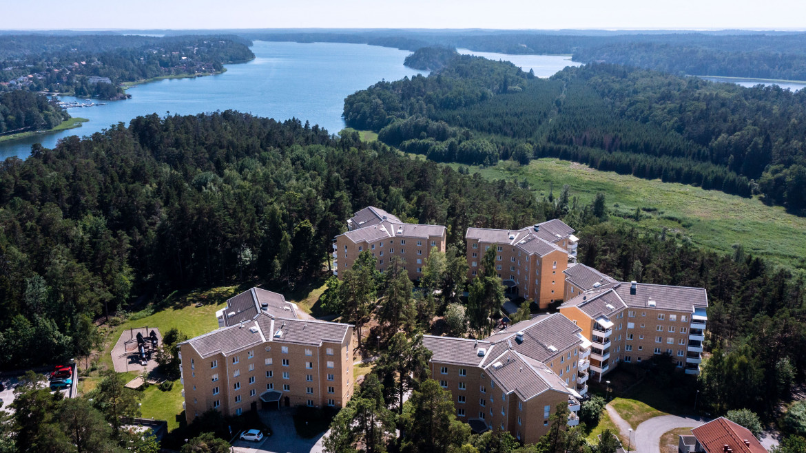 Flerbostadshus på Backluravägen 20 till 28 i Nynäshamn, omgivna av tät skog och belägna nära vatten. Lekplats och parkeringsplatser synliga i förgrunden, med en vy över det natursköna landskapet.