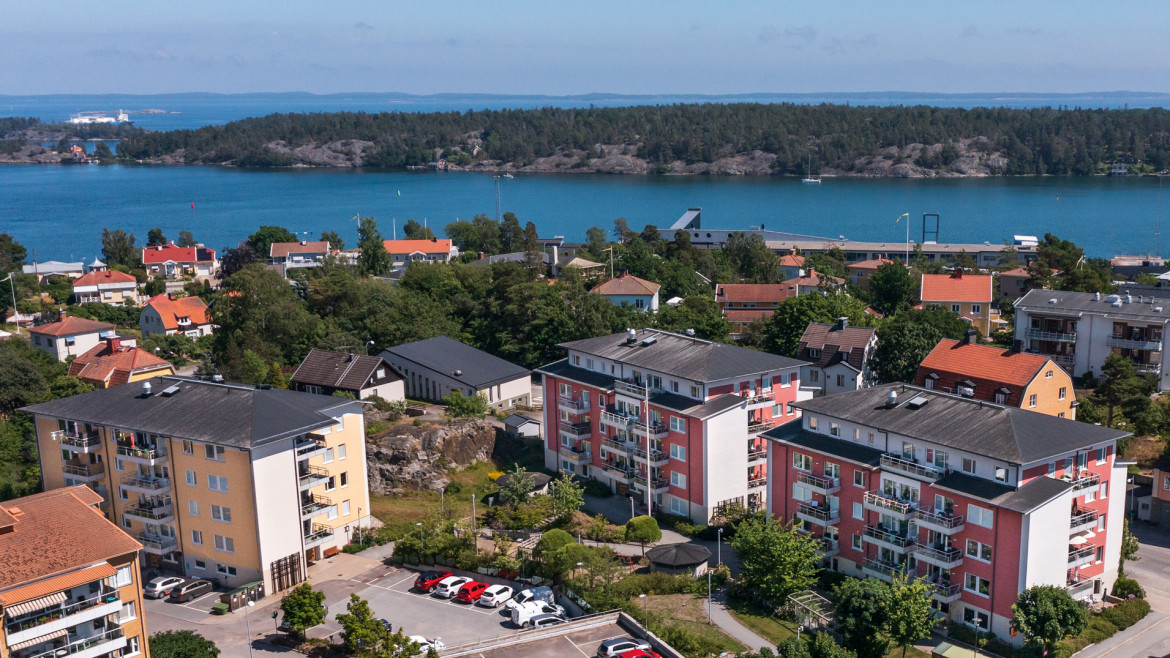 Flygbild över hyreslägenheter på Åkargränd 4, Fredsgatan 19 och 21 i Nynäshamn, med färgglada byggnader och balkonger, omgivna av grönska och med närhet till vatten. Perfekta hyreslägenheter med utsikt över skärgården.