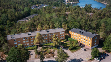 Flygbild över hyreslägenheter på Krokvägen 3 i Nynäshamn, med gula fasader omgivna av skog och vatten. Perfekta hyreslägenheter i en naturskön miljö med närhet till vatten.