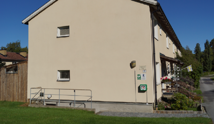 Hjärtstartare monterad på väggen utanför en byggnad på Stora Vika, med en svensk flagga vid entrén och blomsterarrangemang på verandan.