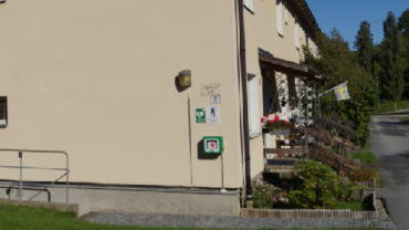 Hjärtstartare monterad på väggen utanför en byggnad på Stora Vika, med en svensk flagga vid entrén och blomsterarrangemang på verandan.