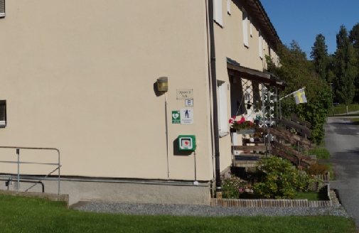Hjärtstartare monterad på väggen utanför en byggnad på Stora Vika, med en svensk flagga vid entrén och blomsterarrangemang på verandan.