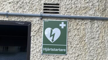 Grön skylt som visar platsen för en hjärtstartare i Heimdal, monterad på en beige, grovputsad vägg. Skylten har ett hjärta med en blixt genom och ett kors i det övre högra hörnet.