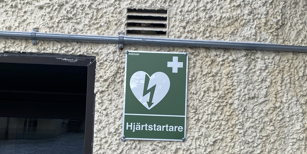 Grön skylt som visar platsen för en hjärtstartare i Heimdal, monterad på en beige, grovputsad vägg. Skylten har ett hjärta med en blixt genom och ett kors i det övre högra hörnet.